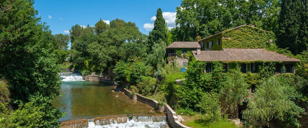 A river flowing through a lush landscape with a stone bridge and a house with ivy-covered walls.
