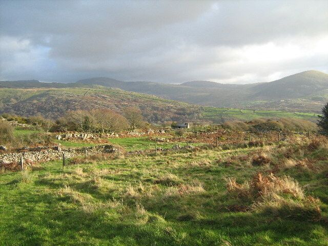 Paisaje rural con colinas ondulantes, un muro de piedra y una edificación distante.