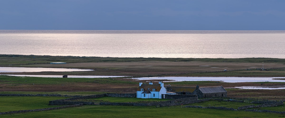 A coastal landscape with a white house surrounded by stone walls, a body of water, and a vast sea in the background.