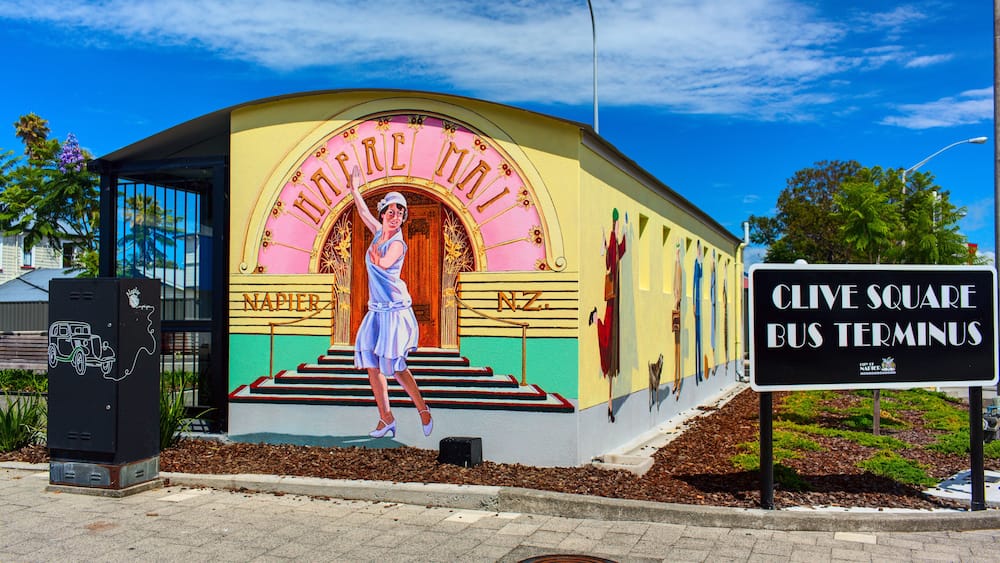 Un bâtiment aux couleurs vives, orné d’une fresque représentant une femme en robe, avec les inscriptions « NAPIER » et « CLIVE SQUARE BUS TERMINUS ».