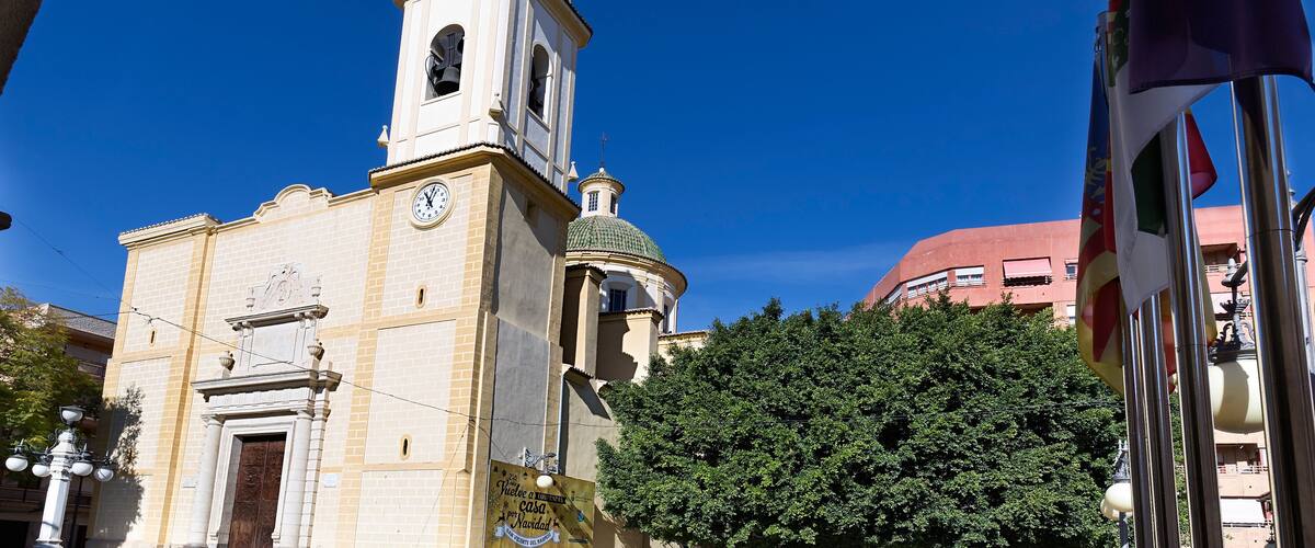 A historic church with a bell tower and a clock, surrounded by trees and flags.