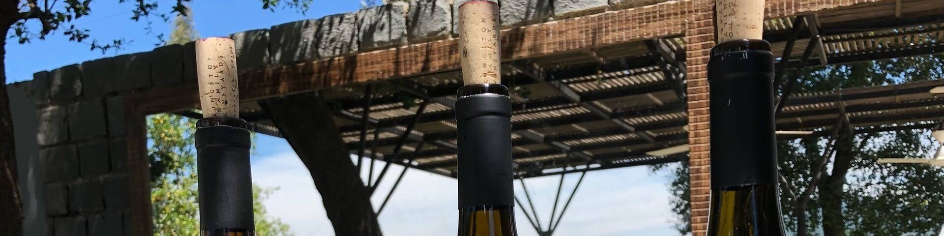 Tres botellas de vino de Kingston Family Vineyards, con etiquetas que indican las variedades 'Torrano' y 'Sucro', colocadas al aire libre sobre una mesa con copas y un dispensador de agua al fondo.
