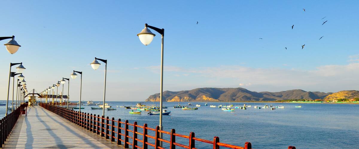A pier with red railings and street lamps, boats on the water, and mountains in the background.