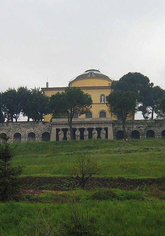 A yellow building with a dome, surrounded by trees and a stone bridge.