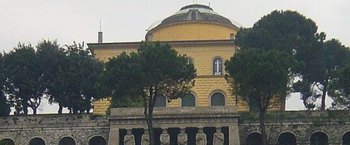 A yellow building with a dome, surrounded by trees and a stone bridge.