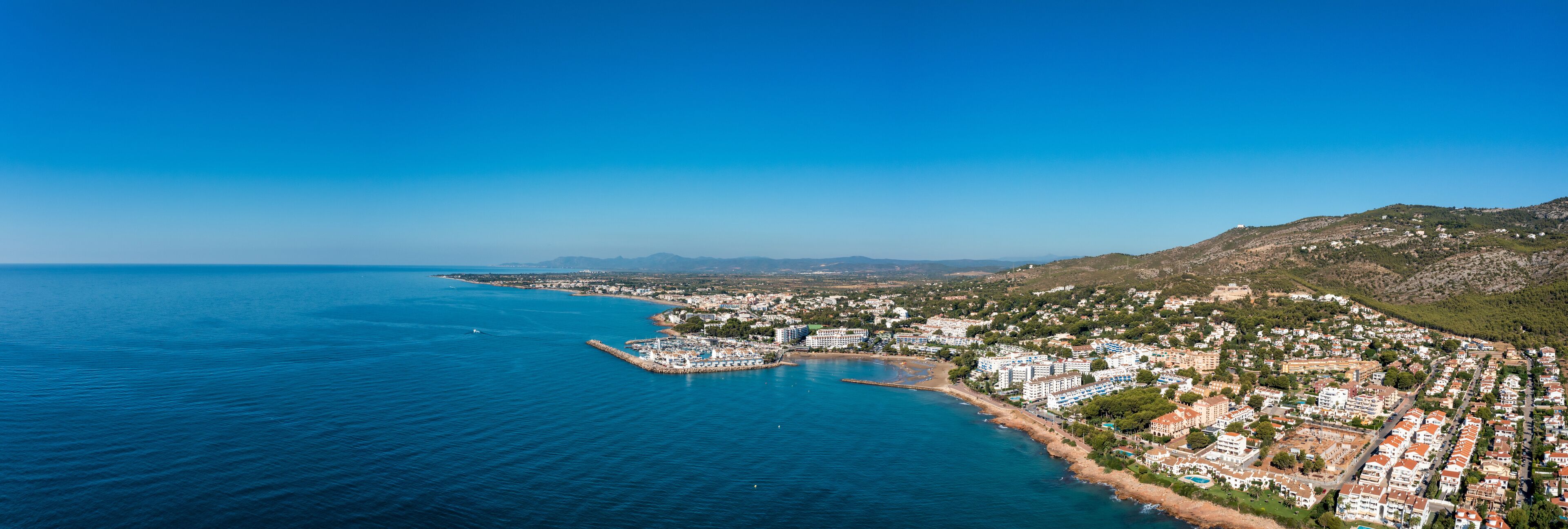 A coastal town with a marina, buildings, and a mountainous backdrop.