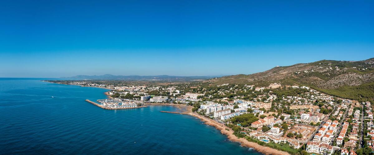 A coastal town with a marina, buildings, and a mountainous backdrop.
