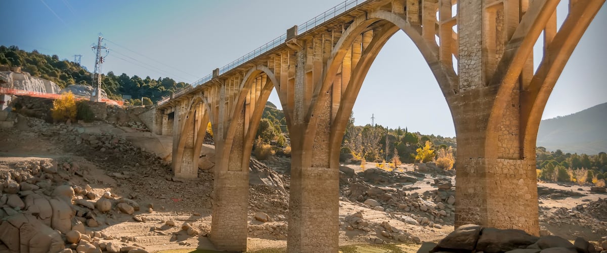 A large stone bridge with multiple arches spanning a river, surrounded by rocky terrain and sparse vegetation.