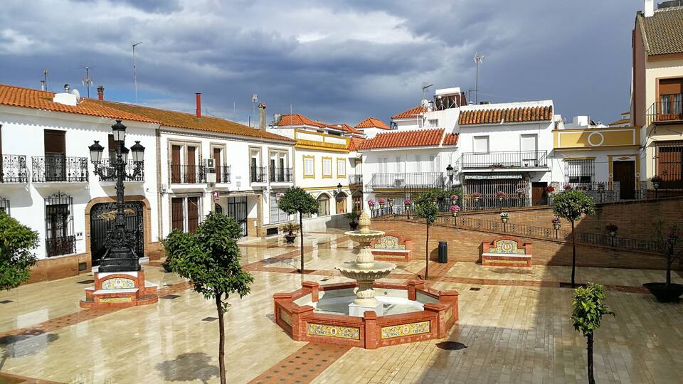 A plaza with tiled flooring, a central fountain, and surrounding buildings with balconies and red-tiled roofs.