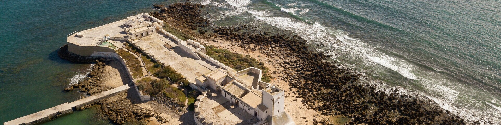 Vista aérea de una fortificación costera con un litoral rocoso circundante y aguas azules cristalinas.