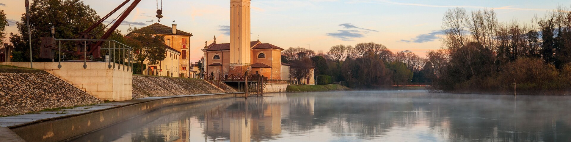 Un río tranquilo que refleja un edificio histórico con una torre alta, rodeado de árboles y un muelle de madera.