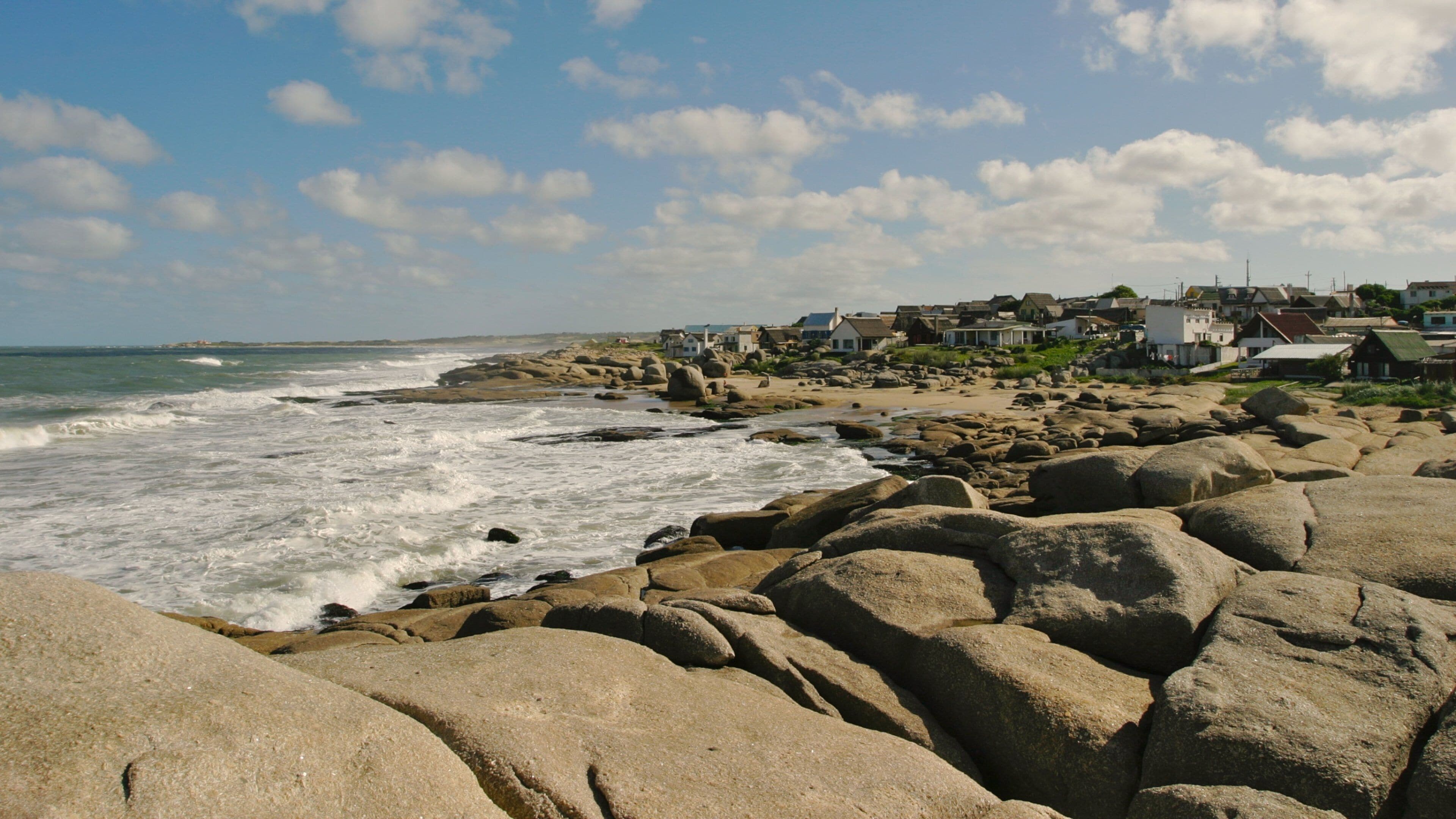 Una costa rocosa con olas rompiendo contra las rocas, un pequeño pueblo costero con casas, y un cielo azul despejado con nubes.