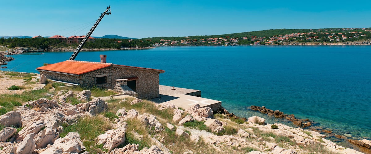 A coastal village with a stone building on a rocky shore, a clear blue sea, and a distant hilly landscape.