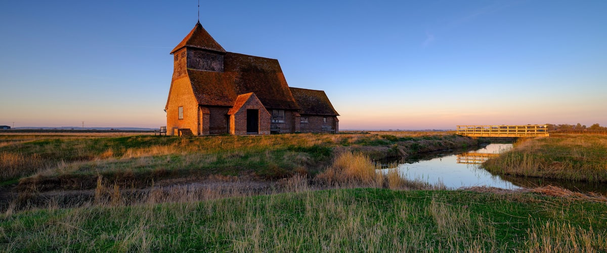 A brick church with a steep roof and a tower, surrounded by a marshy landscape and a small wooden bridge.