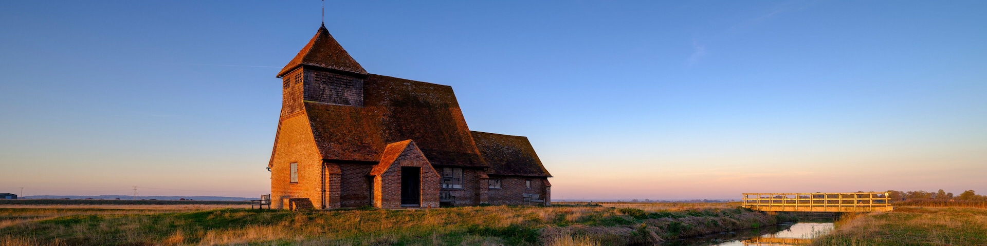 A brick church with a steep roof and a tower, surrounded by a marshy landscape and a small wooden bridge.