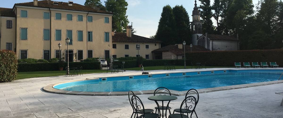 A swimming pool with lounge chairs and a table, surrounded by a paved area and buildings in the background.