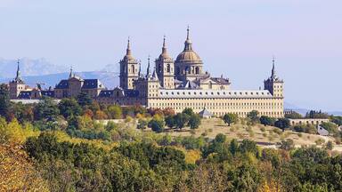A large, historic building with multiple domes and towers, surrounded by trees and mountains in the background.