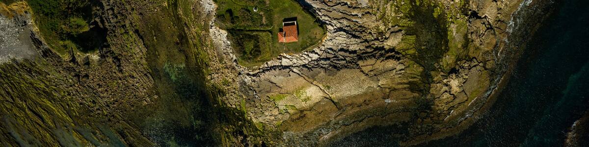 Vue aérienne d’une zone côtière avec un petit bâtiment entouré d’un terrain rocheux et d’une végétation verdoyante.