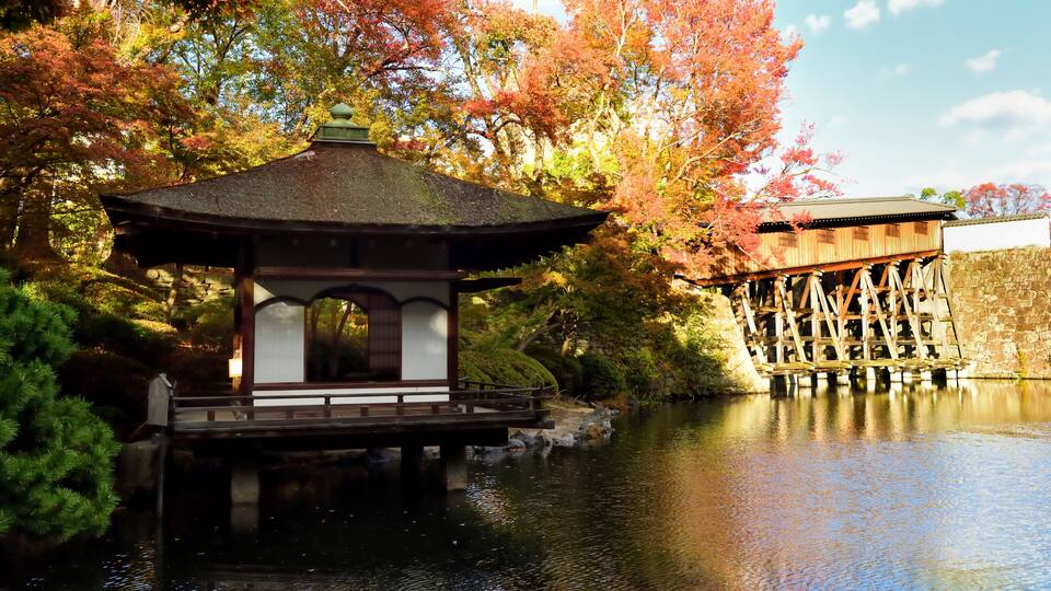 A traditional Japanese pavilion by a calm river, surrounded by autumn foliage and a wooden waterwheel structure.
