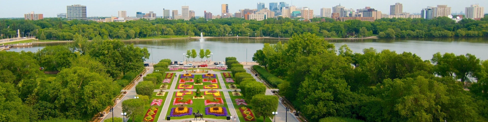 A city park with a lake, surrounded by trees and a parking lot with cars.