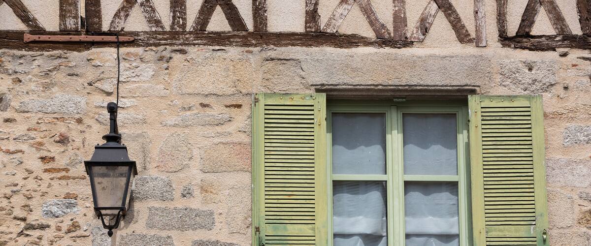 A stone building with a window featuring green shutters and a white sailcloth hanging below.