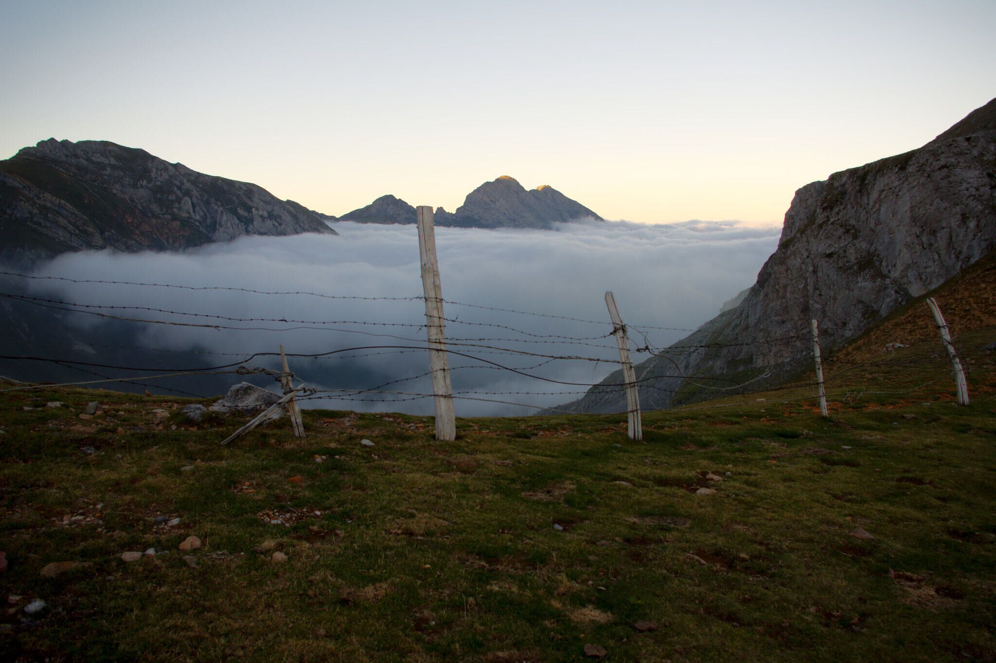 A mountainous landscape with a barbed wire fence, rocky cliffs, and a valley shrouded in clouds.