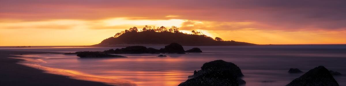 Panorama long exposure of an orange sunrise across a seascape scene of rocks looking out towards the silhouette of Green Island at Cunjurong Point, New South Wales Australia
