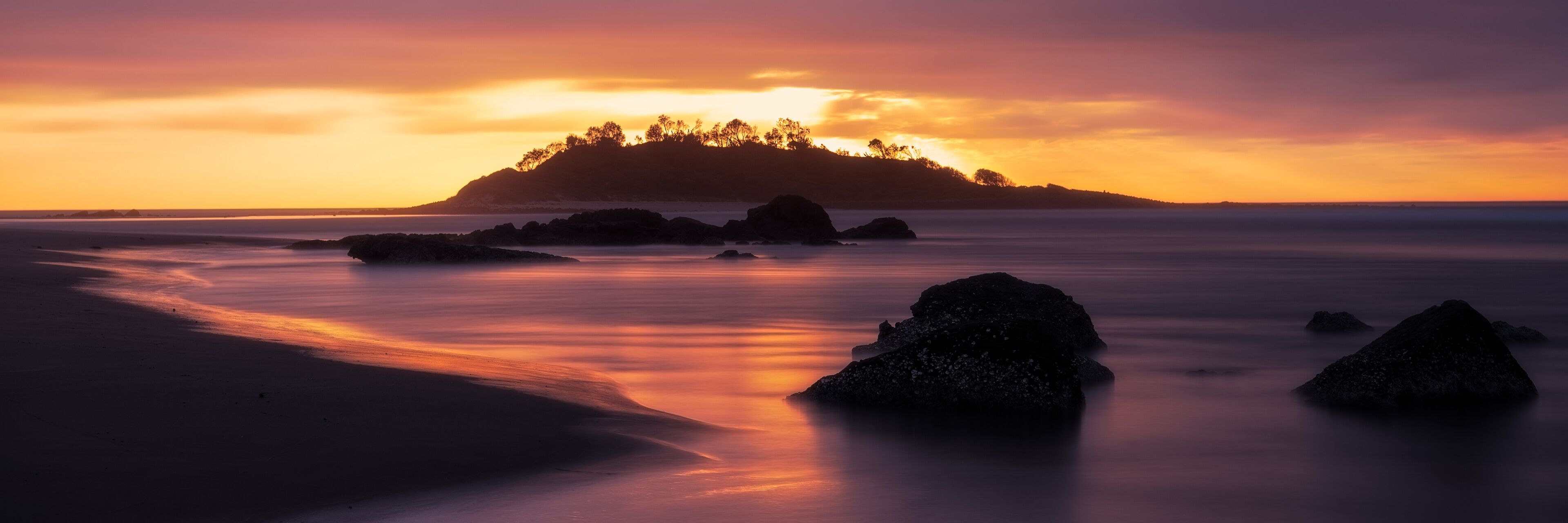 A sunset over a beach with rocks and a forested island in the distance.