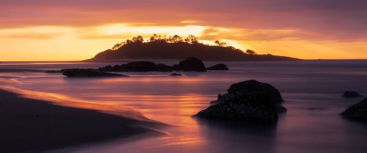 A sunset over a beach with rocks and a forested island in the distance.