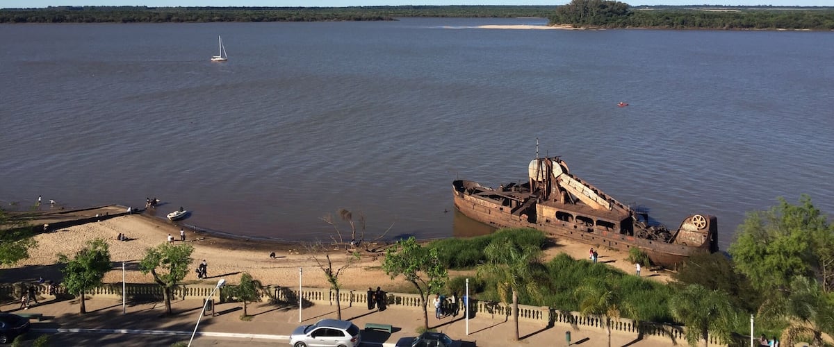 A large body of water with a rusted shipwreck on the shore, a sandy beach, and a clear blue sky.