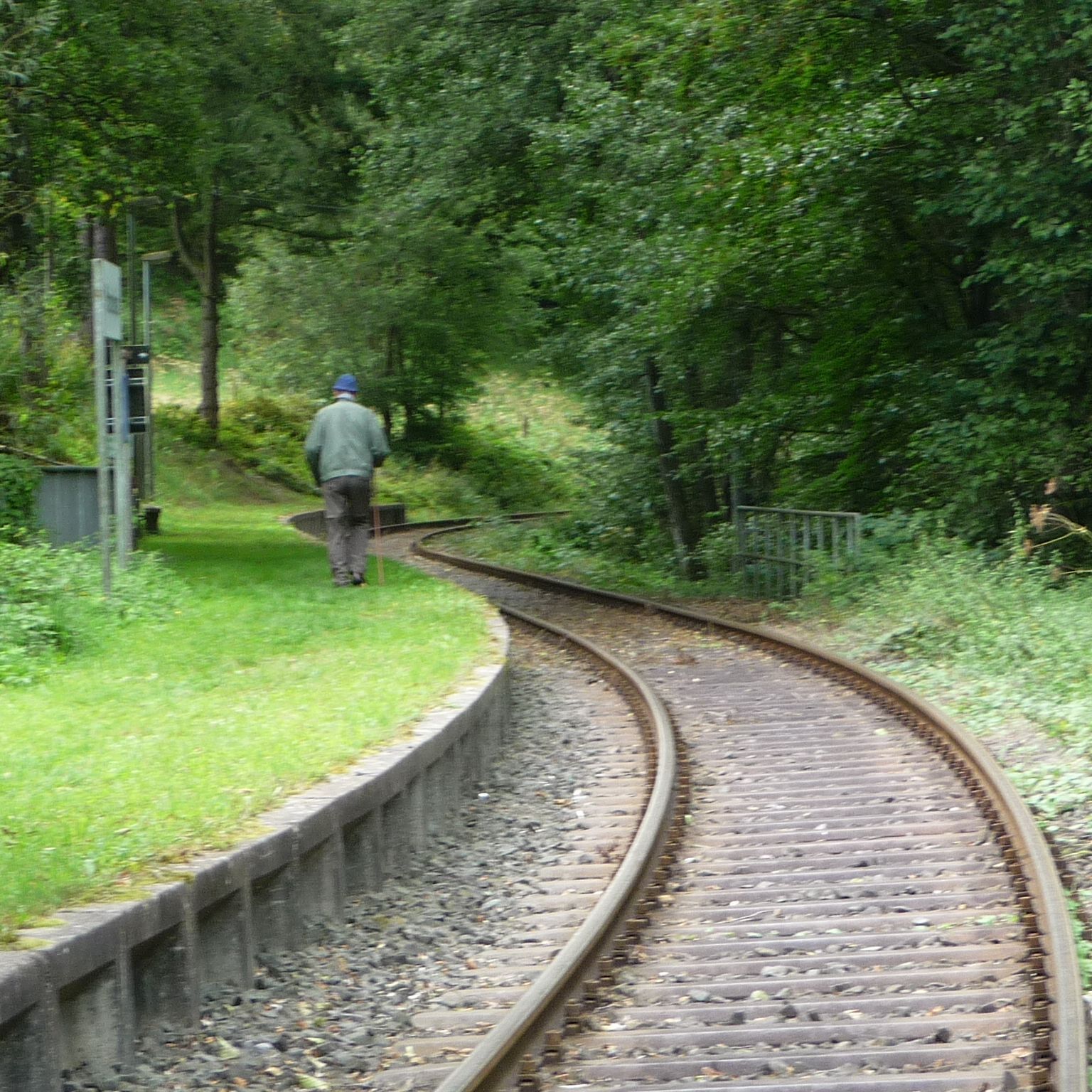 A railway track curving through a wooded area with a person walking on the grass beside it.
