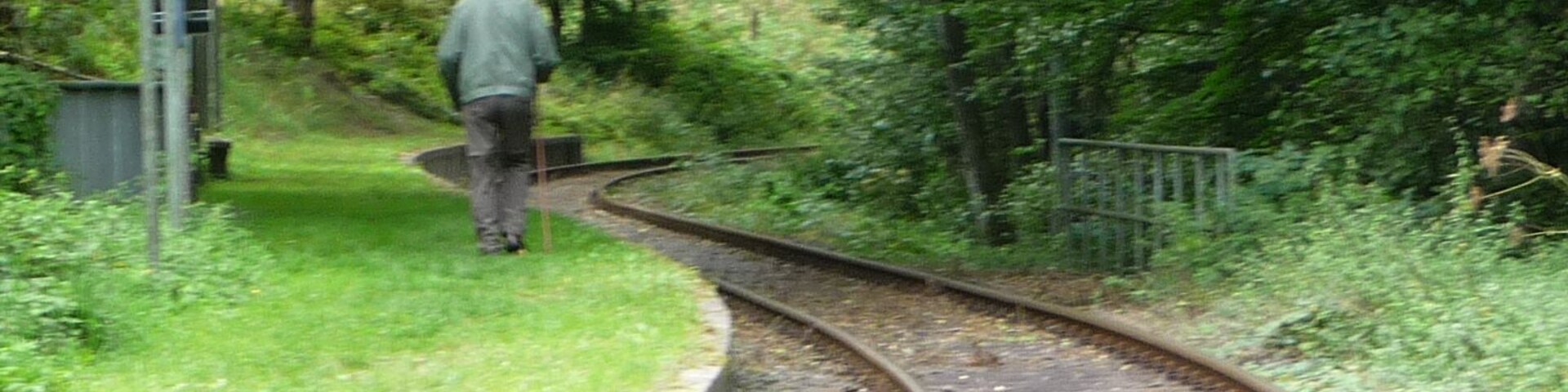 A railway track curving through a wooded area with a person walking on the grass beside it.