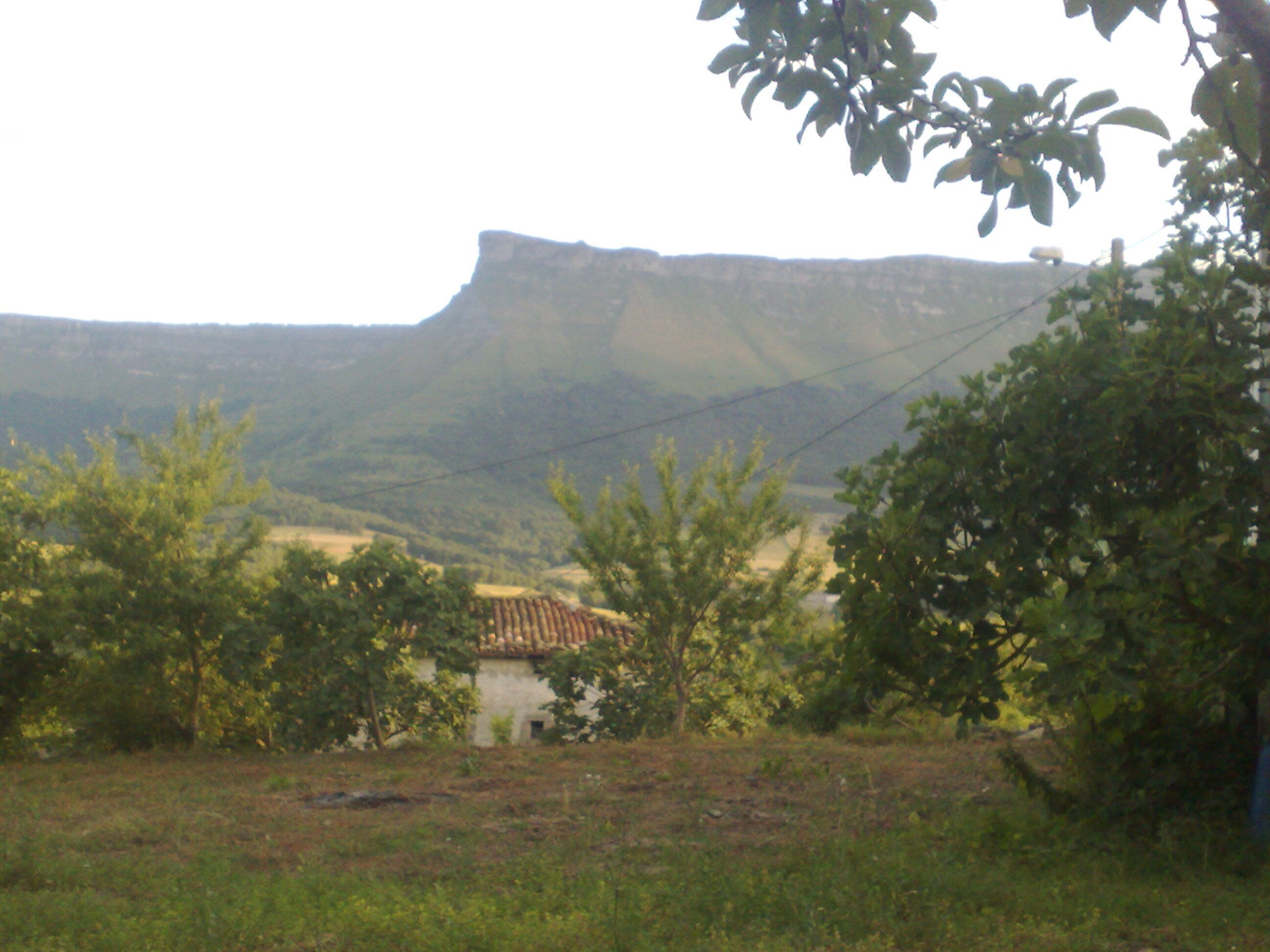 A mountainous landscape with a prominent cliff, trees, and a building in the foreground.