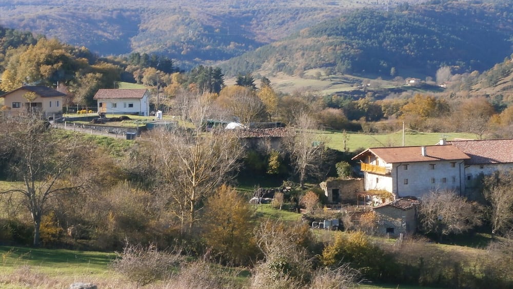 Ländliche Landschaft mit Häusern, einer Brücke und sanften Hügeln im Hintergrund.
