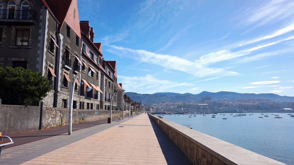 A waterfront promenade with historic buildings, a clear sky, and boats in the water.