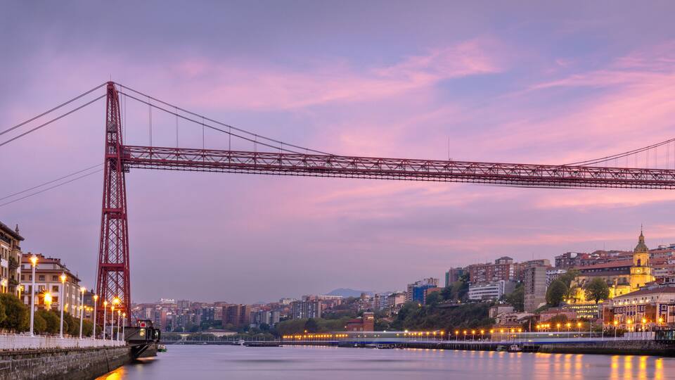 A red bridge spanning a river with city buildings on either side.