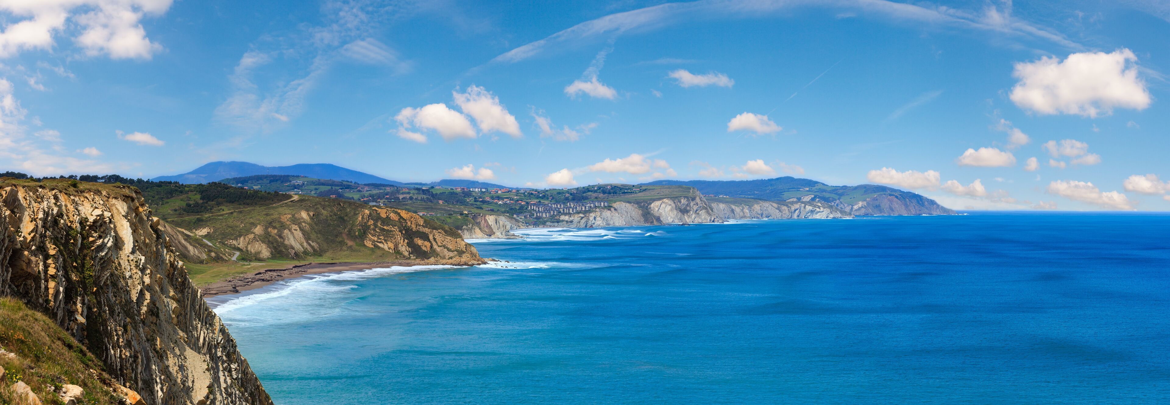 A coastal landscape with cliffs, a beach, and distant mountains under a clear sky.