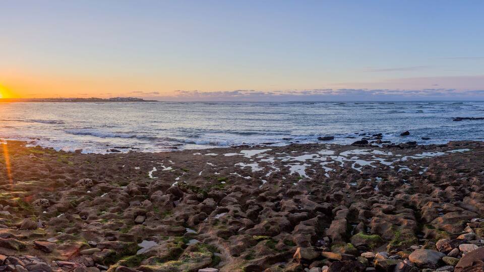 A rocky shoreline at sunset with the sun low on the horizon.