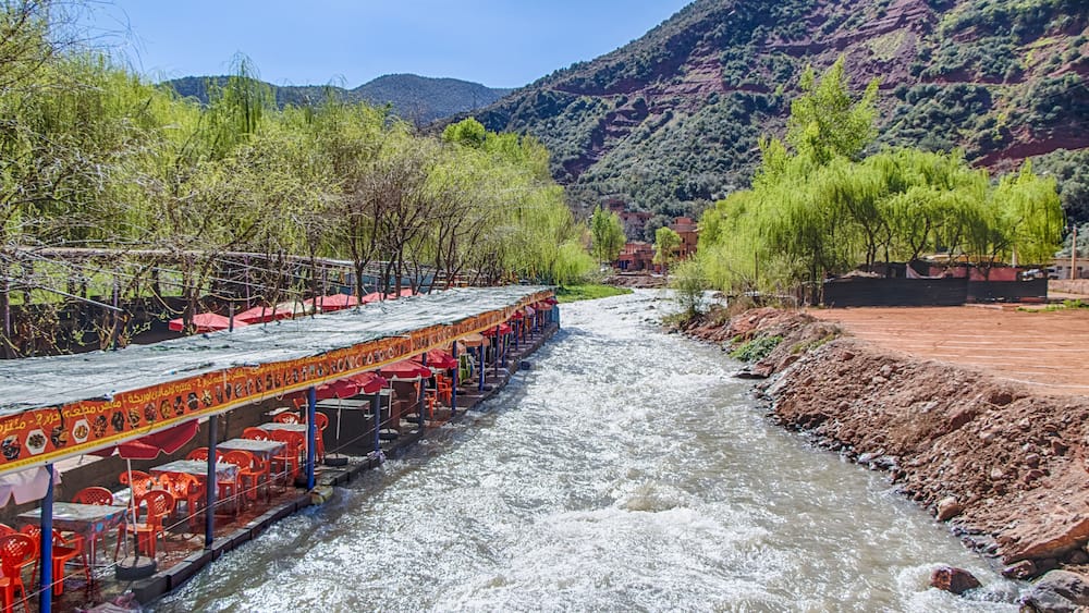 A river with a boat restaurant on the left, surrounded by greenery and mountains in the background.