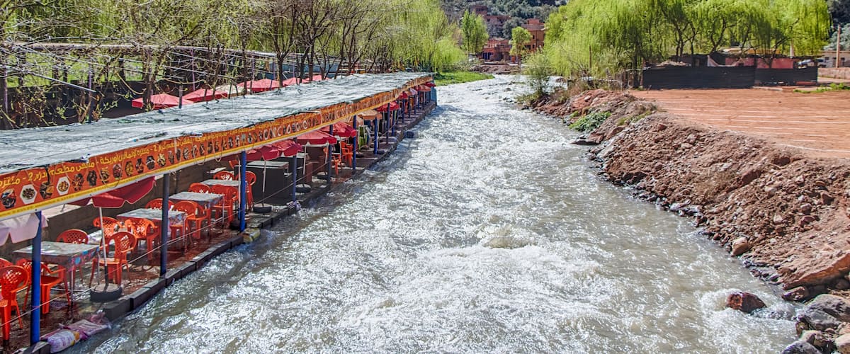 A river with a boat restaurant on the left, surrounded by greenery and mountains in the background.