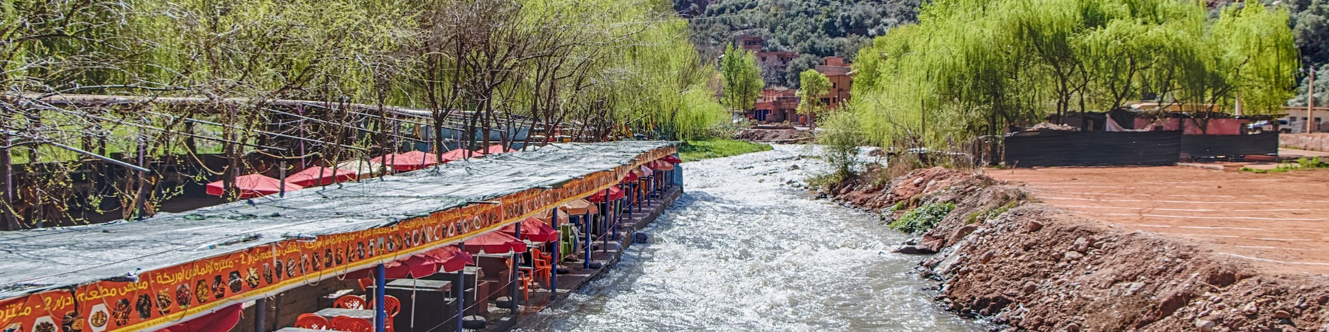 A river with a boat restaurant on the left, surrounded by greenery and mountains in the background.