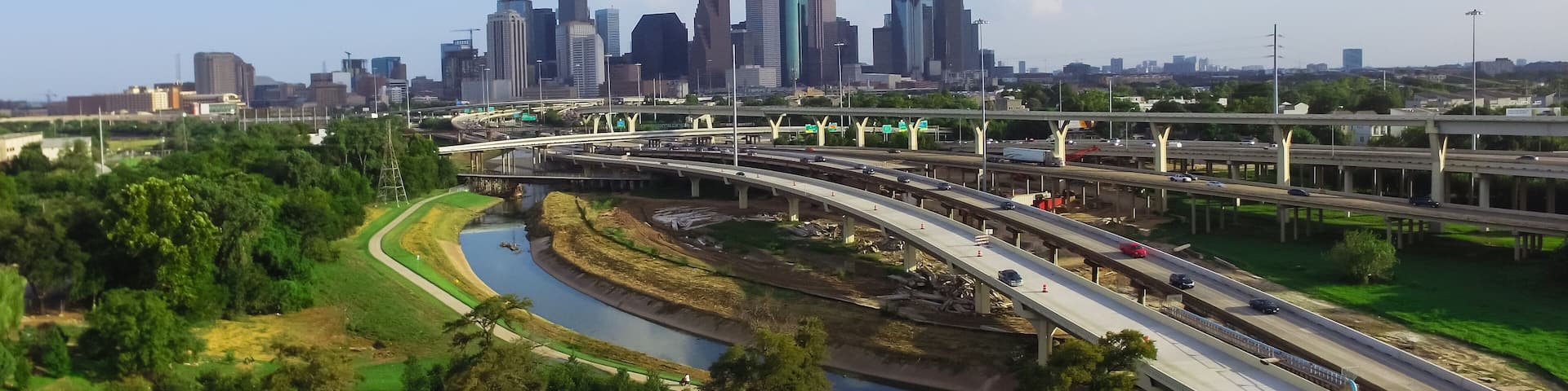 A city skyline with modern skyscrapers, a highway with multiple lanes, and a river curving through a green park area.