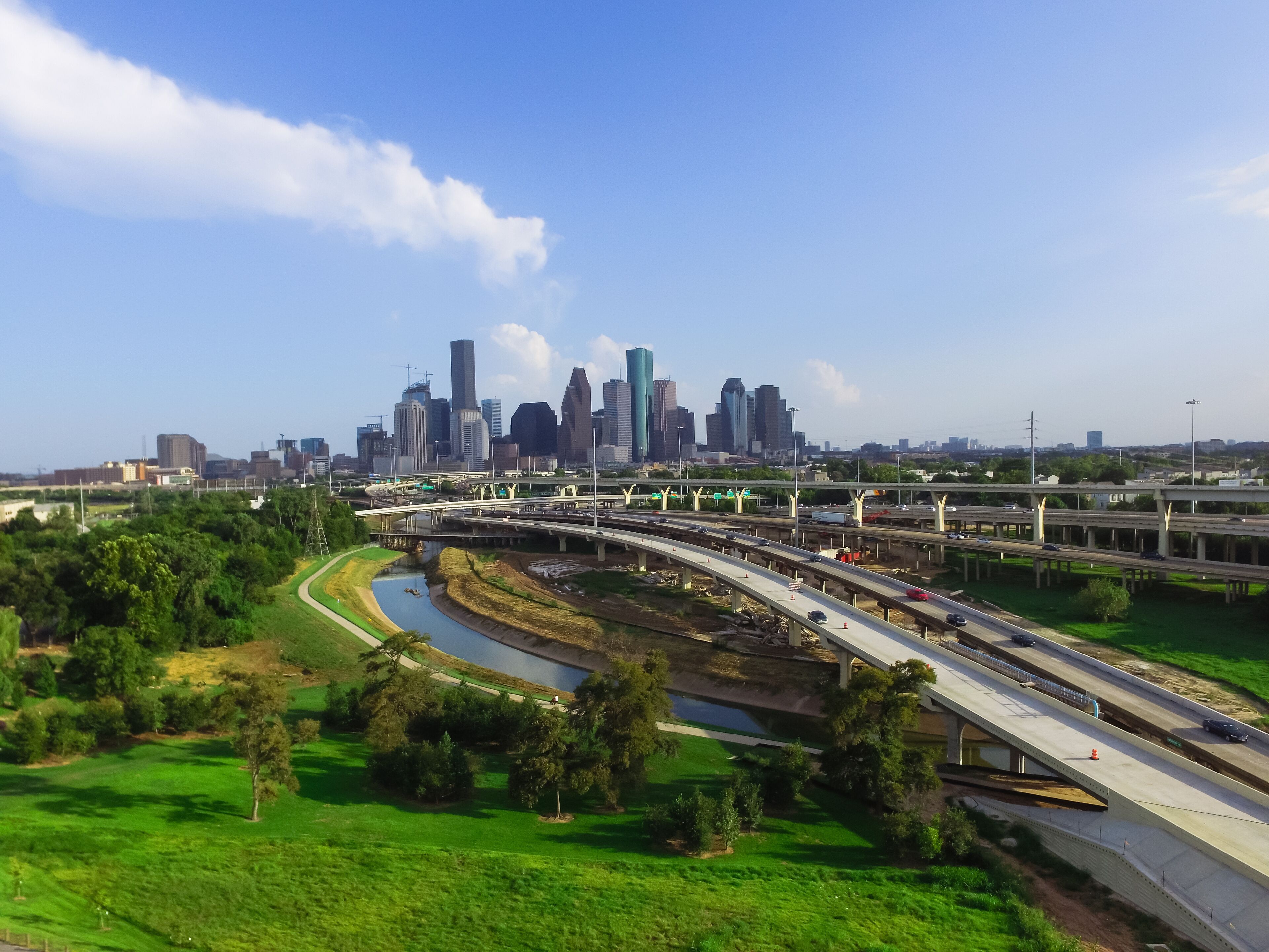 A city skyline with modern skyscrapers, a highway with multiple lanes, and a river curving through a green park area.