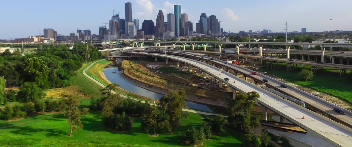 A city skyline with modern skyscrapers, a highway with multiple lanes, and a river curving through a green park area.
