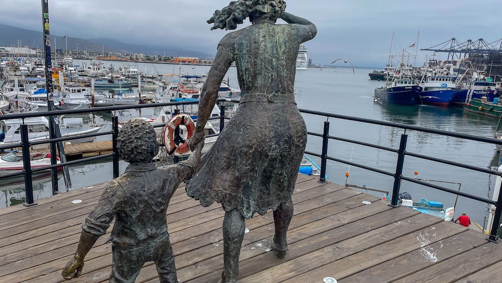 Eine Statue einer Frau und eines Kindes mit Blick auf einen Yachthafen, wo Boote und eine Brücke in der Ferne liegen.