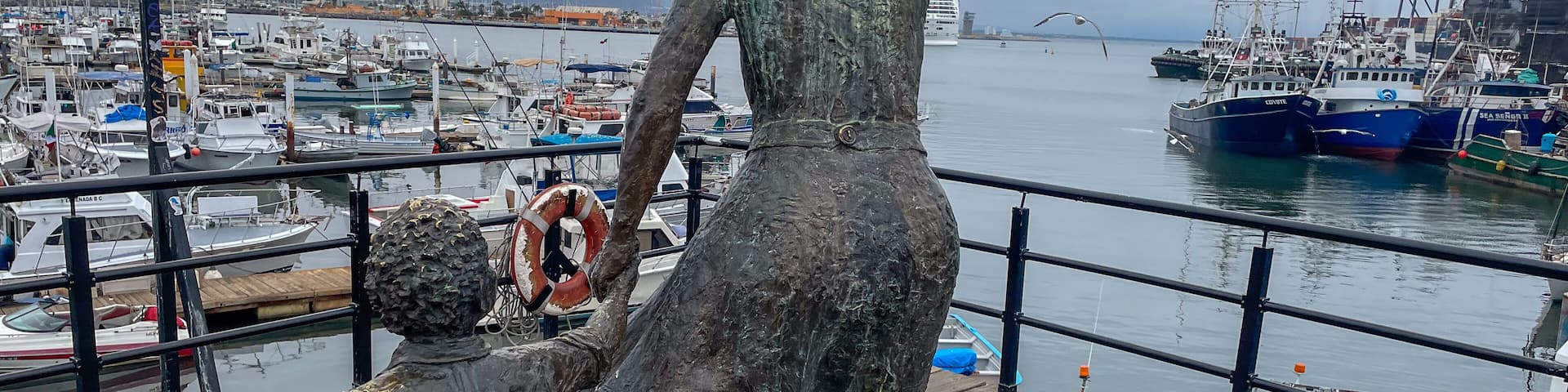 A statue of a woman and child overlooking a marina with boats and a distant bridge.