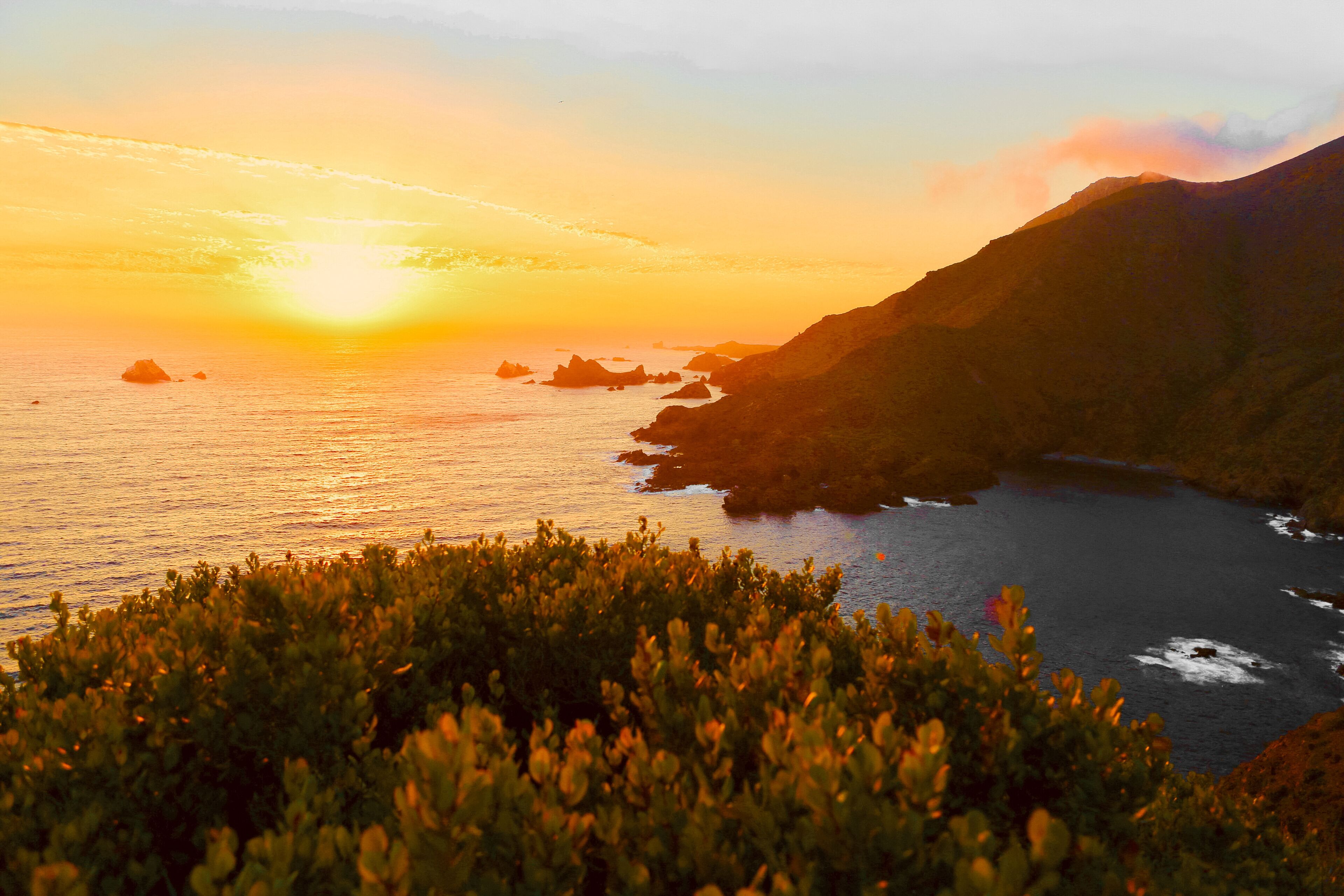 A coastal sunset with a mountain range and dense vegetation in the foreground.