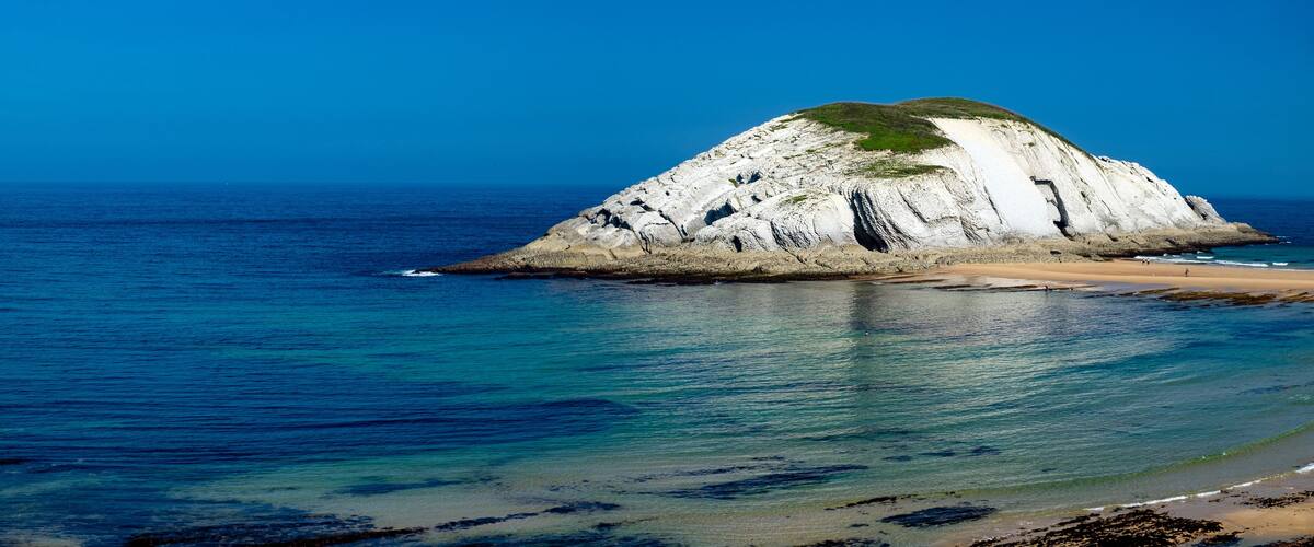A coastal landscape with a prominent white rock formation surrounded by clear blue waters and a sandy beach.