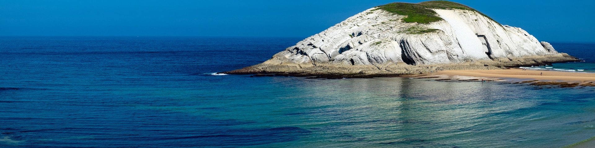 A coastal landscape with a prominent white rock formation surrounded by clear blue waters and a sandy beach.