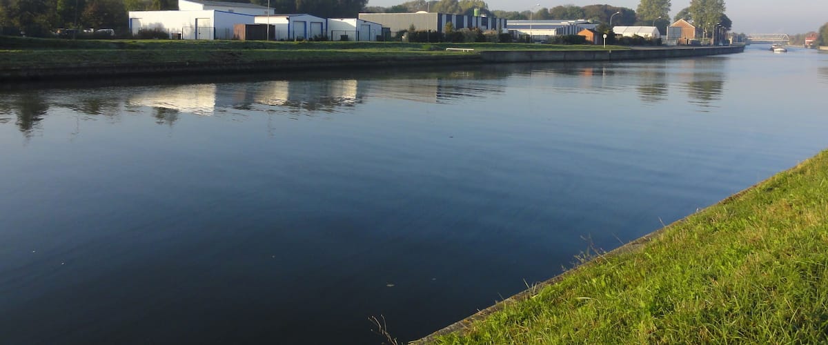 A calm river with industrial buildings and a bridge in the background.